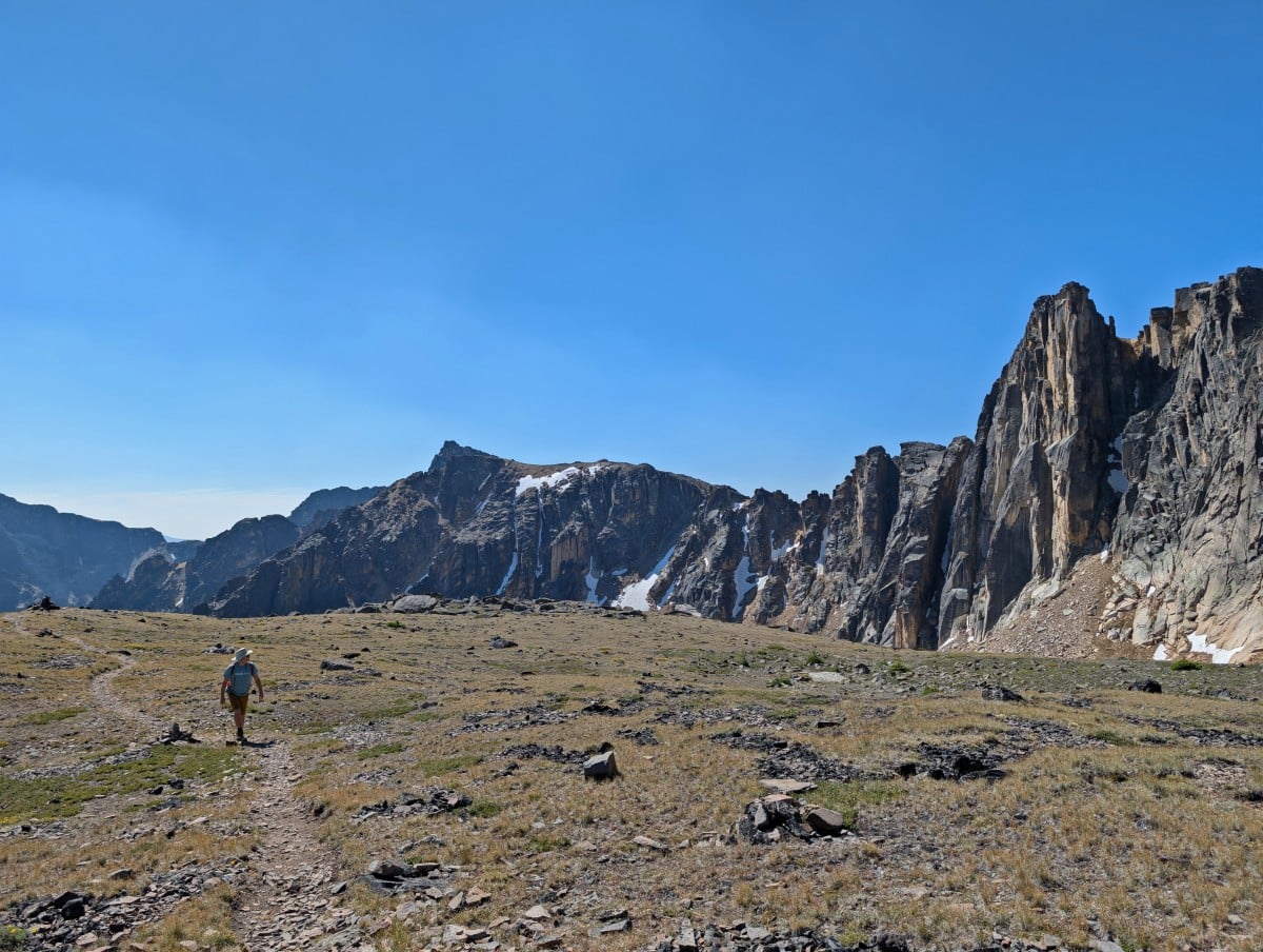 A hiker walks towards camera on dirt path, through open alpine scenery with rock wall of mountains on right side