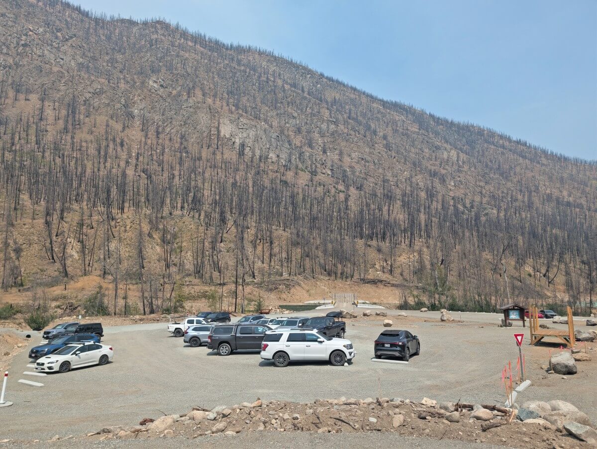 View of Cathedral Lakes Lodge parking area with large dirt area, parked vehicles and mountain slope behind