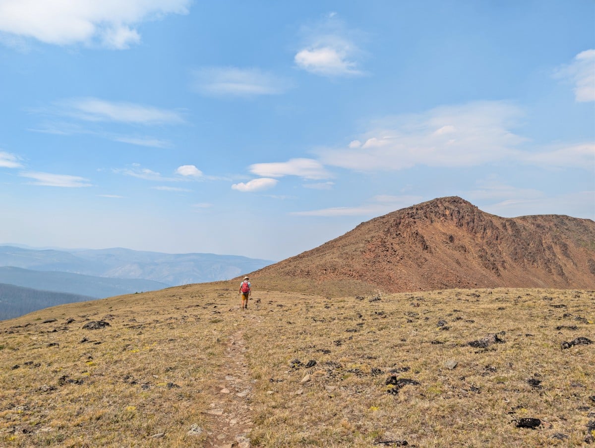 Back view of hiker heading rowards red tinged Red Mountain, surrounded by open alpine scenery
